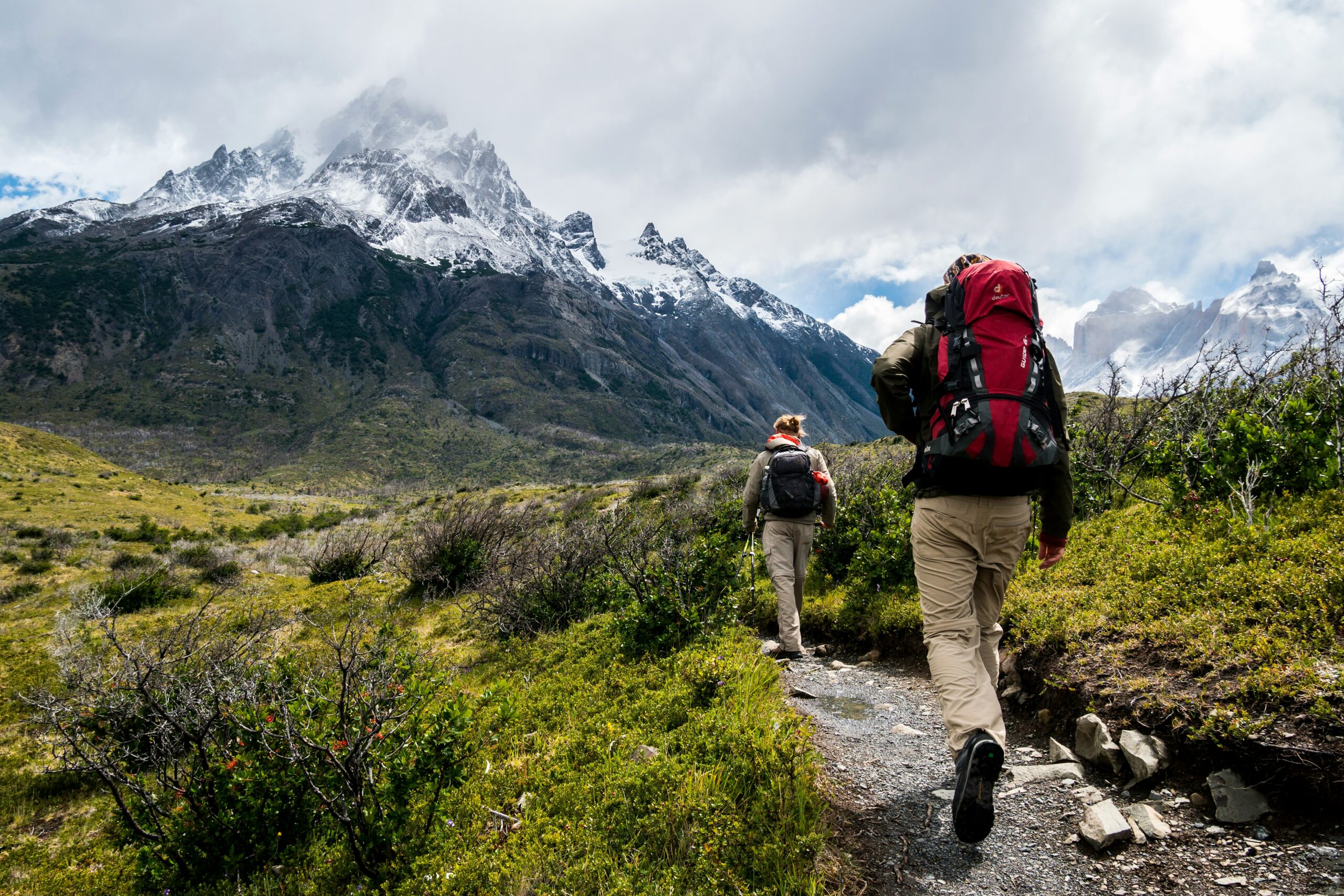 Two hikers trekking on a mountain trail towards snow-capped peaks.